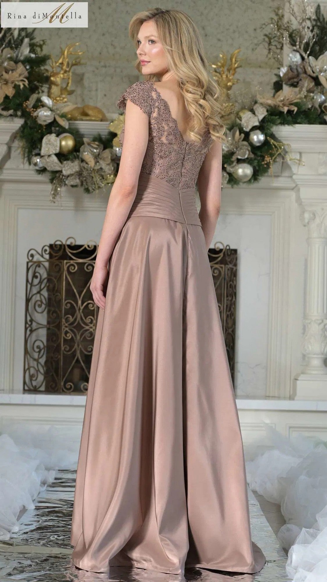 A woman with long blonde hair models the Rina Di Montella RD3327, a long formal dress with a lace-embroidered bodice and satin champagne finish, standing before a holiday-adorned mantle.